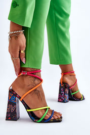 Woman wearing Eliore high heel buckled sandals with colorful straps and patterned wide heel, paired with green pants.