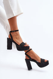 Woman wearing black textile platform stiletto sandals with adjustable buckle, paired with white pants, showcasing stylish footwear.