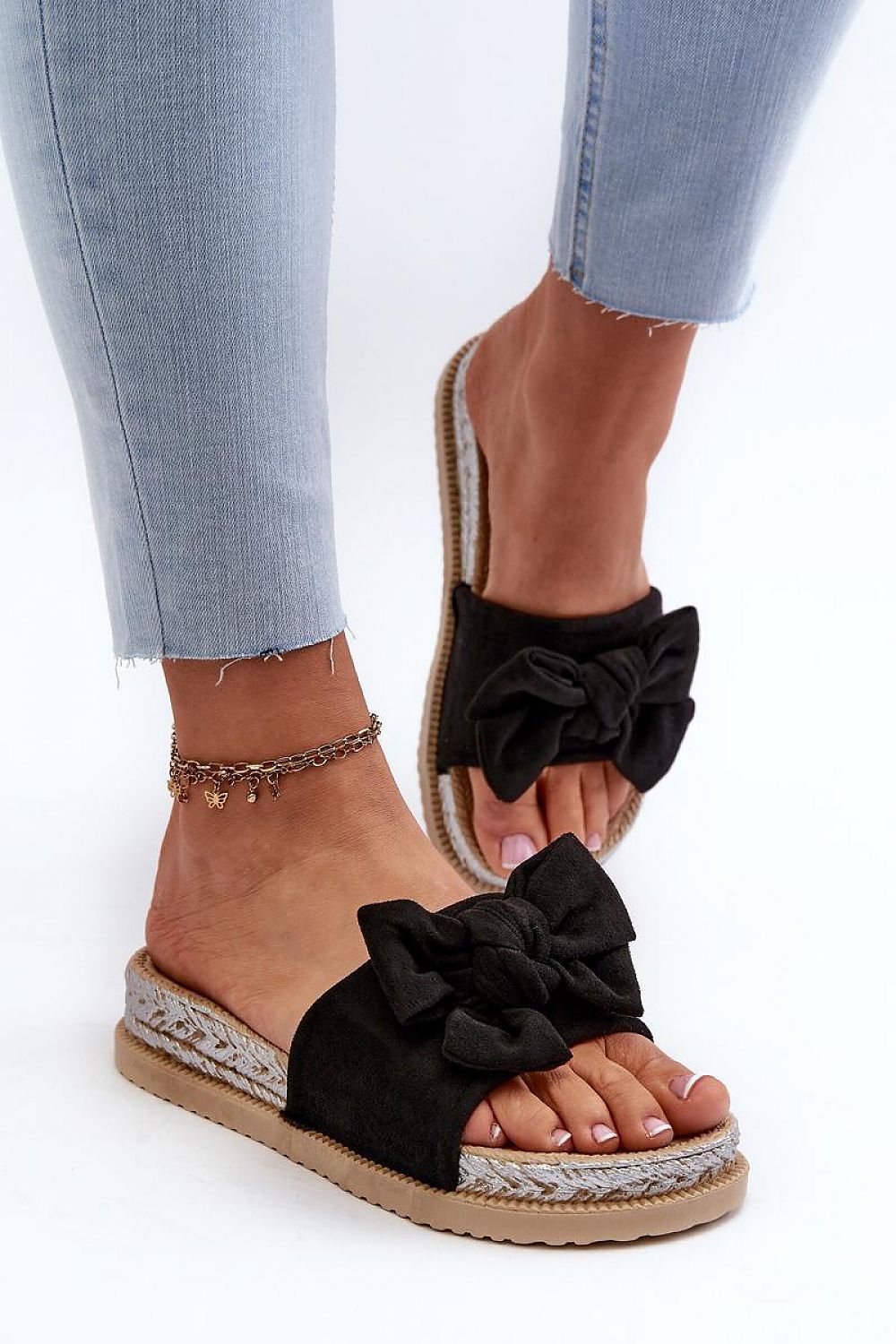 Black Alinara Suede Knot Platform Slides with bow design on model's feet, wearing light jeans and anklet.