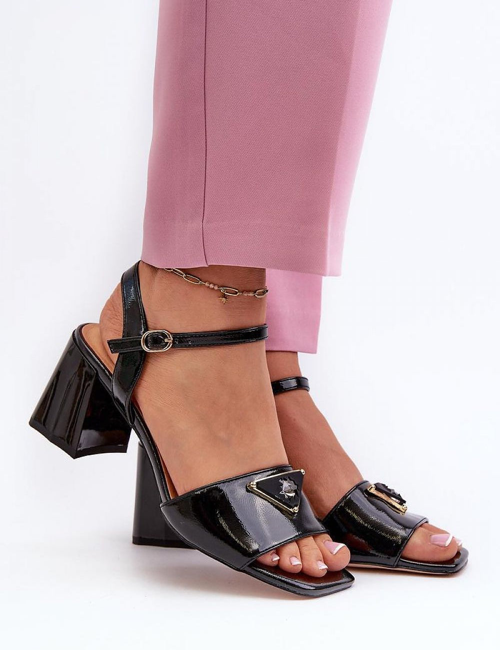 Glossy block heel sandals with ankle straps and decorative accents, worn with pink pants. Lacquered artificial leather upper, chic design.