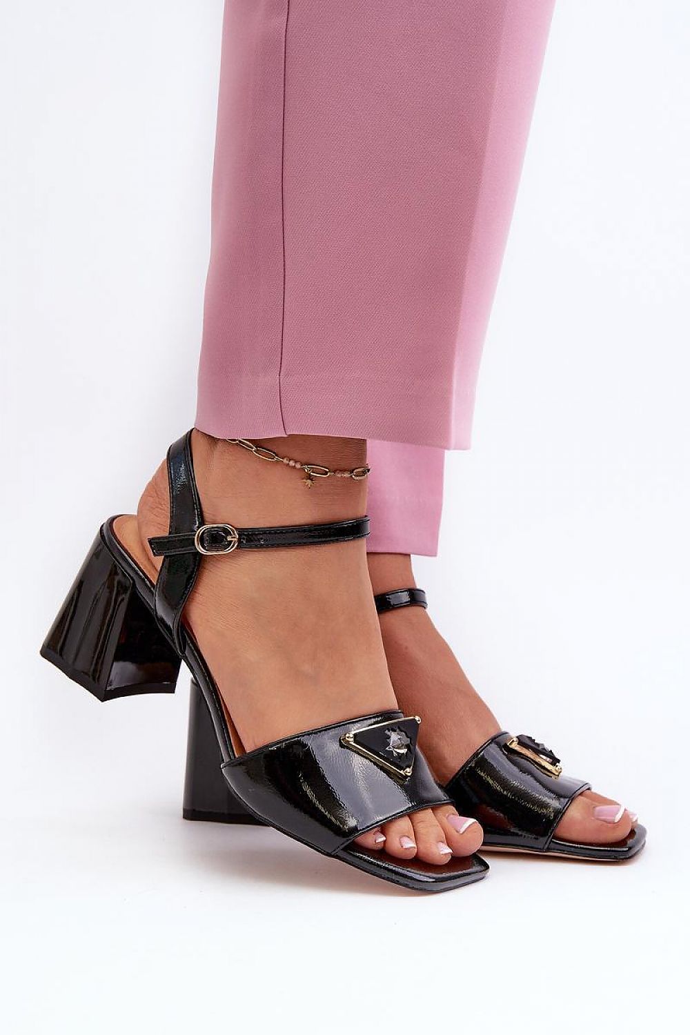 Glossy block heel sandals with ankle straps and decorative accents, worn with pink pants. Lacquered artificial leather upper, chic design.
