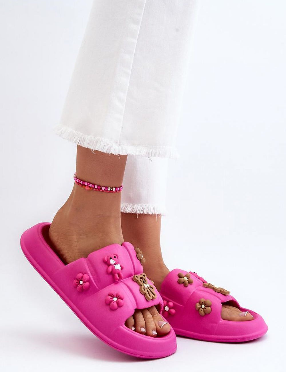Pink Valessa foam platform summer slides with broad strap, whimsical accents, soft plush material, paired with white pants and beaded anklet.