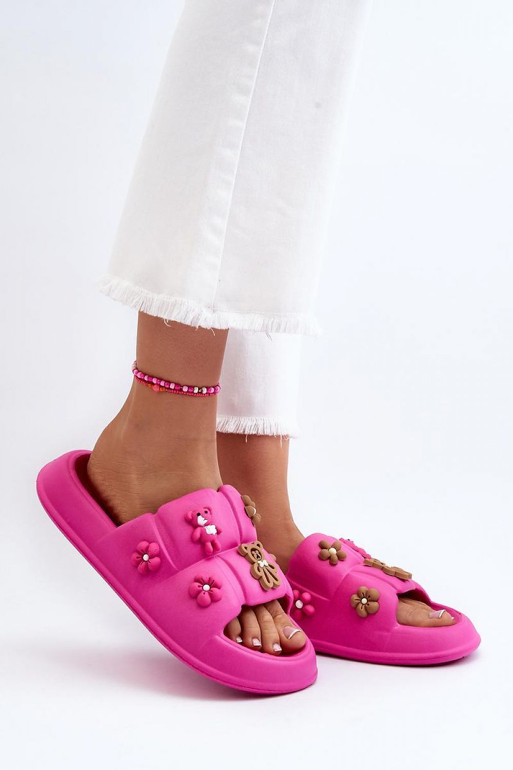 Pink Valessa foam platform summer slides with broad strap, whimsical accents, soft plush material, paired with white pants and beaded anklet.