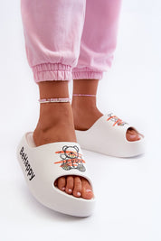 Person wearing white slip-on shoes with teddy bear print and 'Be Happy' text, paired with pink pants and anklet.