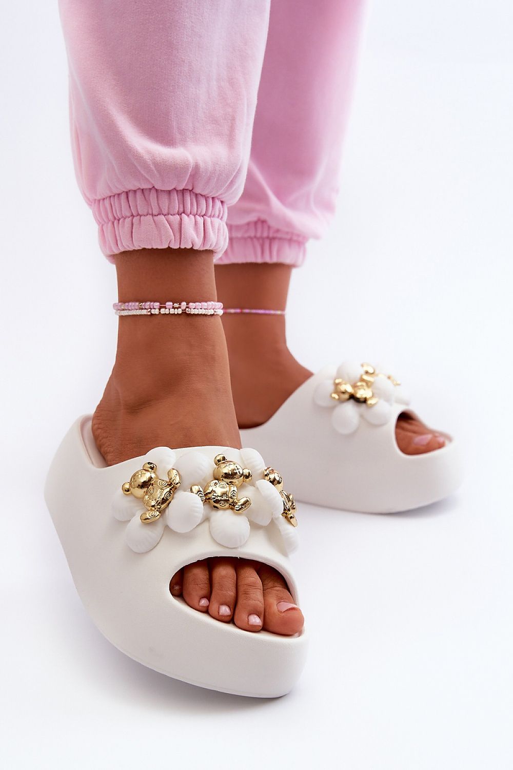 Person wearing white embellished platform sandals with decorative straps, paired with pink pants and a pink anklet, on a white background.