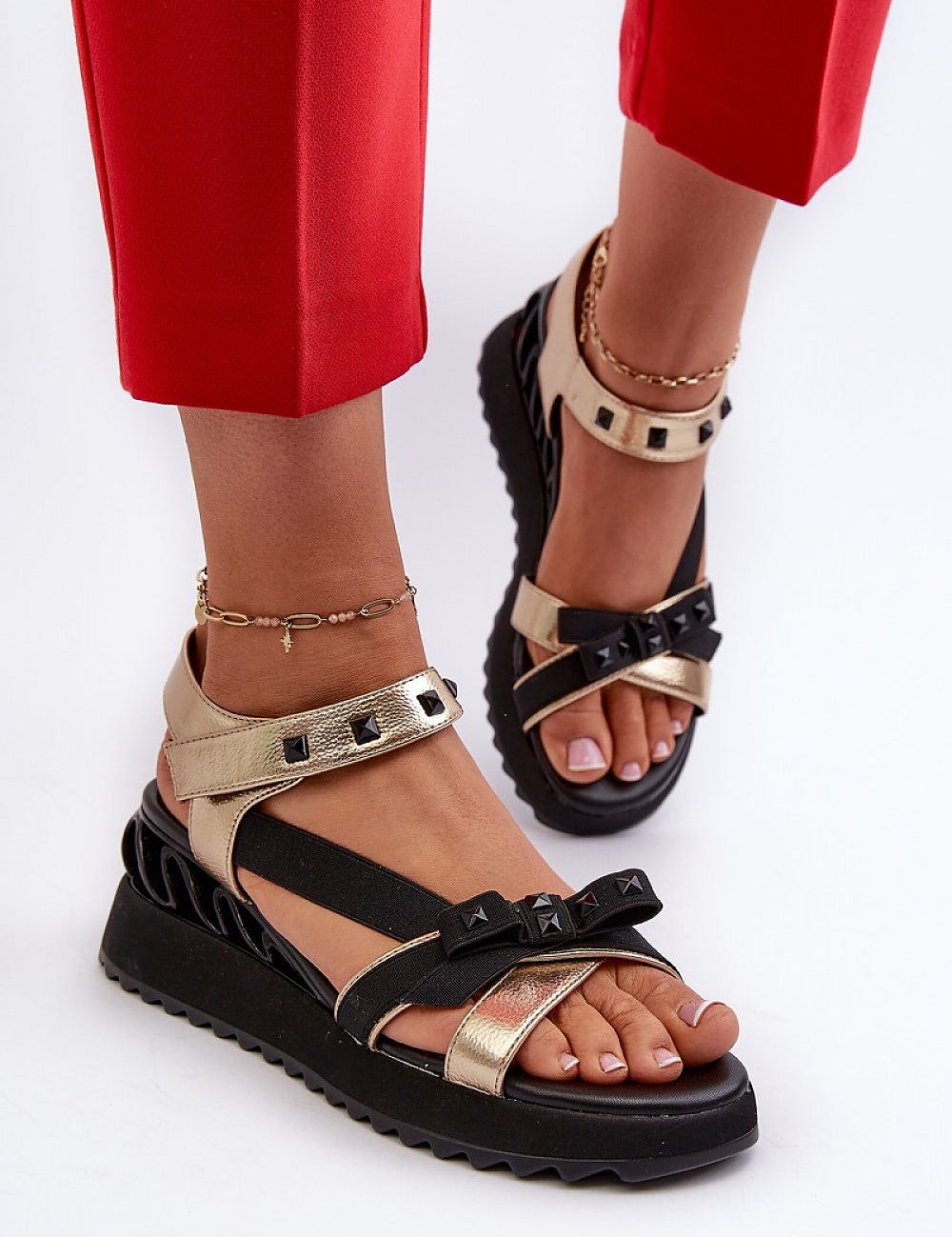 Gold and black bow wedge sandals with ankle straps, shown on feet with red pants, featuring embellished details and platform heels.