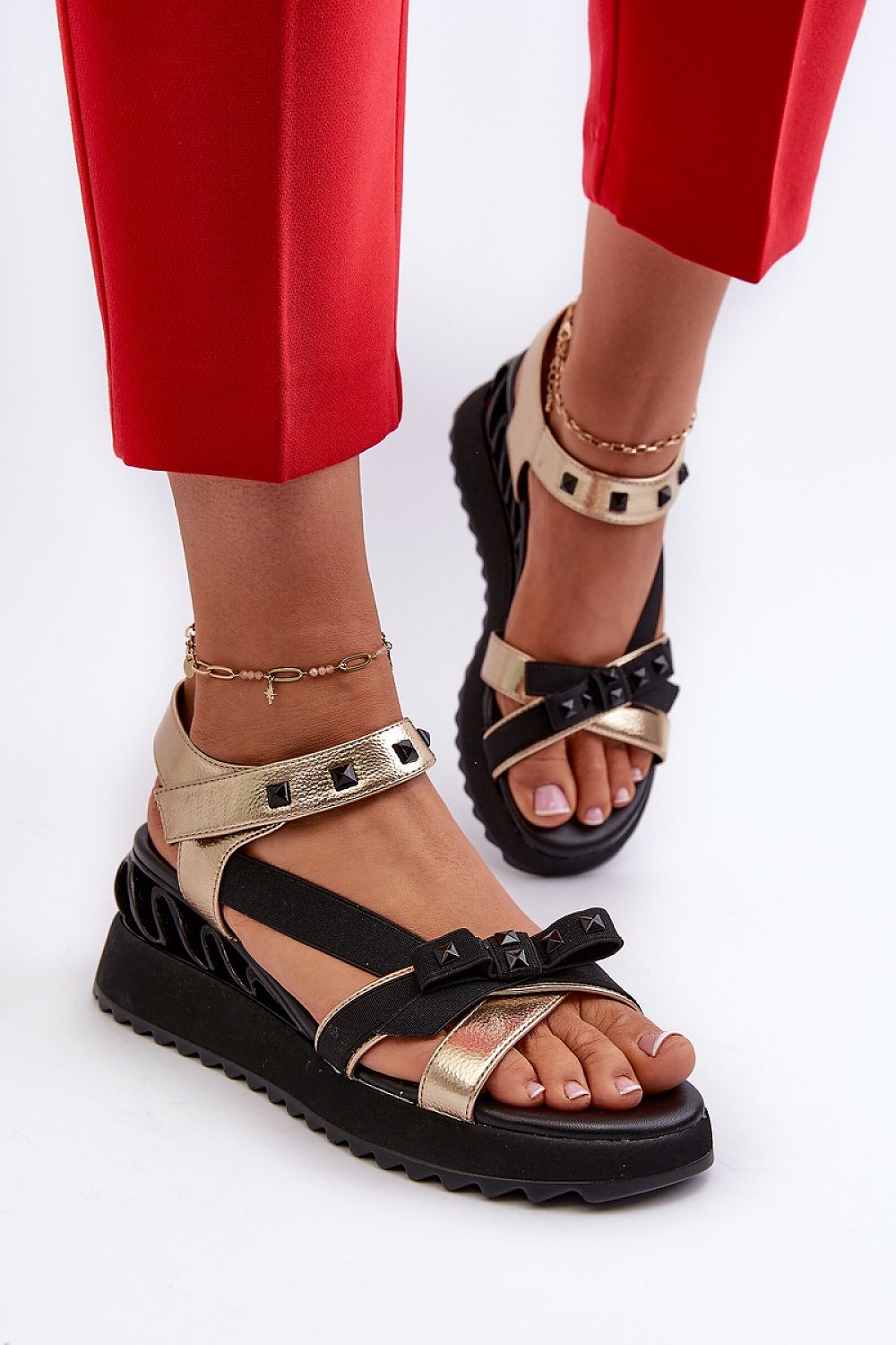 Gold and black bow wedge sandals with ankle straps, shown on feet with red pants, featuring embellished details and platform heels.