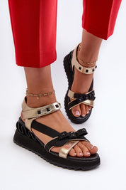 Gold and black bow wedge sandals with ankle straps, shown on feet with red pants, featuring embellished details and platform heels.