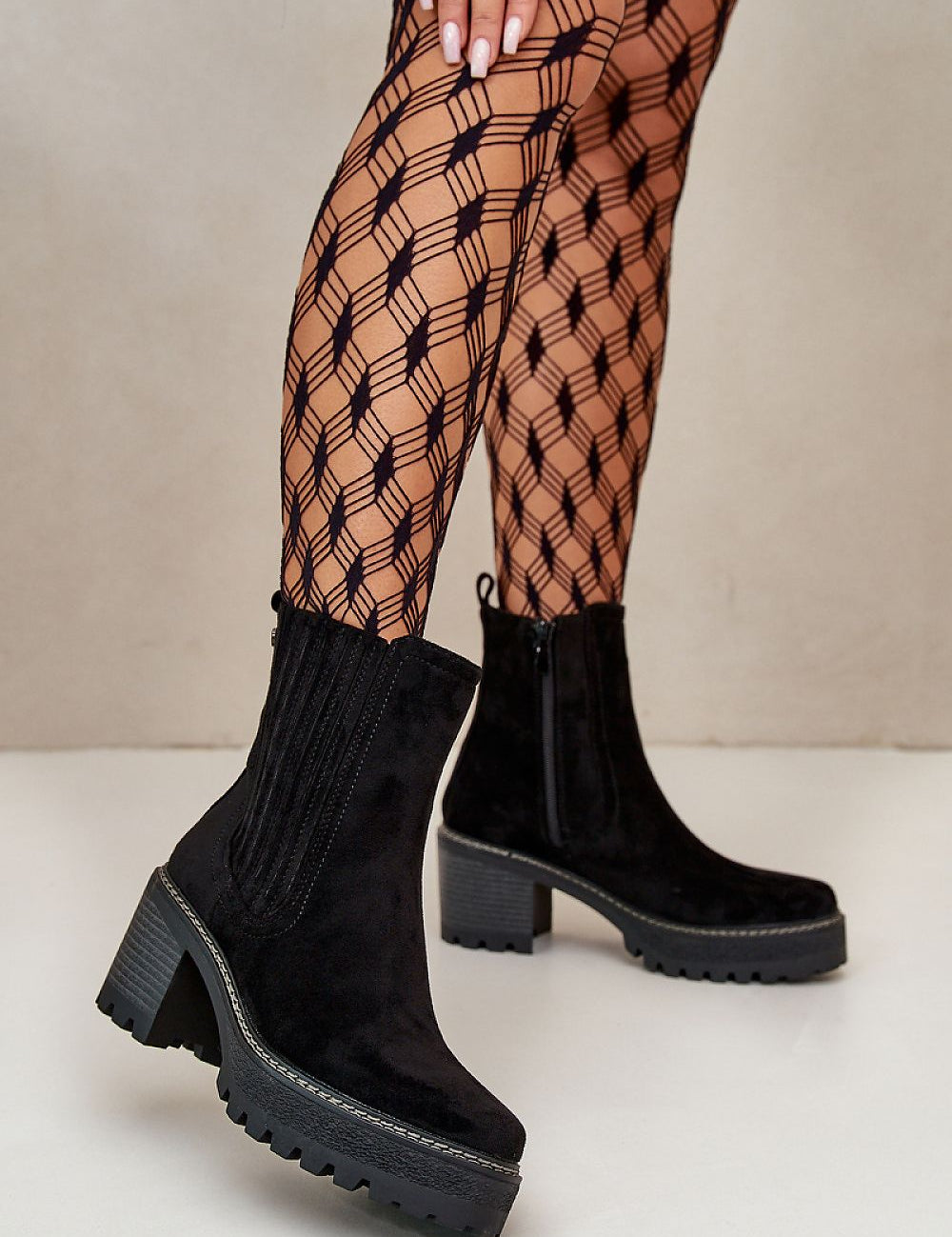 Woman wearing black suede-finish ankle boots with block heel and patterned tights, showcasing stylish footwear in a modern setting.