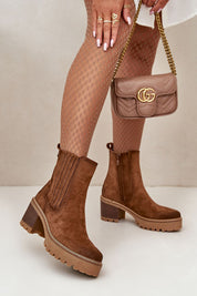 Brown suede-finish ankle boots with block heels and side zipper, paired with a stylish tan handbag and patterned tights.
