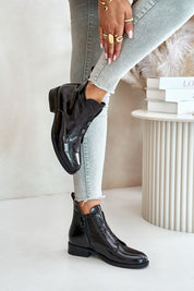 Black leather ankle boots with side zippers worn by a model in gray jeans, standing by a white decorative table.