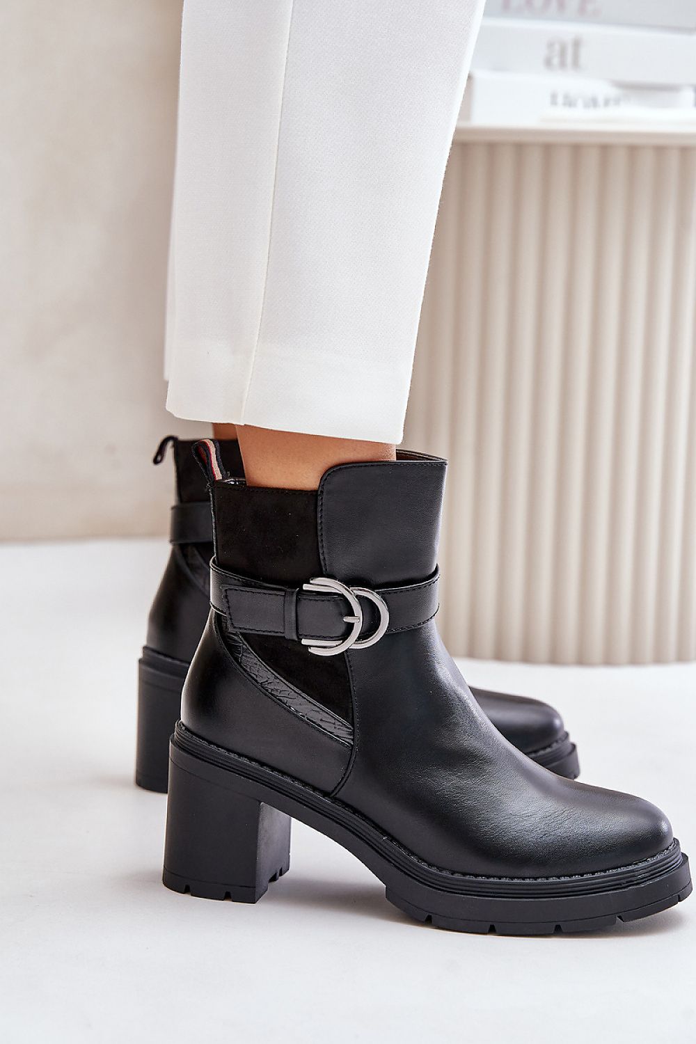 Woman wearing Velona heeled winter boots in black eco leather with gold buckle strap and post heel, paired with white pants.