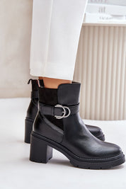 Woman wearing Velona heeled winter boots in black eco leather with gold buckle strap and post heel, paired with white pants.