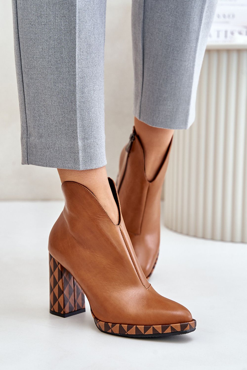 Brown leather ankle boots with geometric heels, styled with gray pants, featuring almond-shaped toe and side zipper.