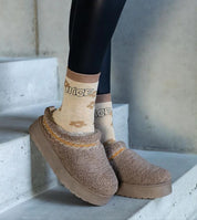 Cozy shearling slip-on slippers with embroidery worn on stairs, paired with tan embroidered socks and black leggings for comfort.