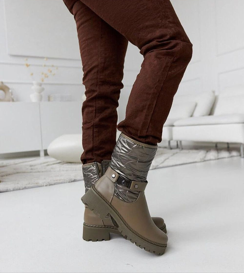 Person wearing Renvara Strap Detail Ankle Boots with insulated design and decorative strap, styled with brown pants in a modern interior.