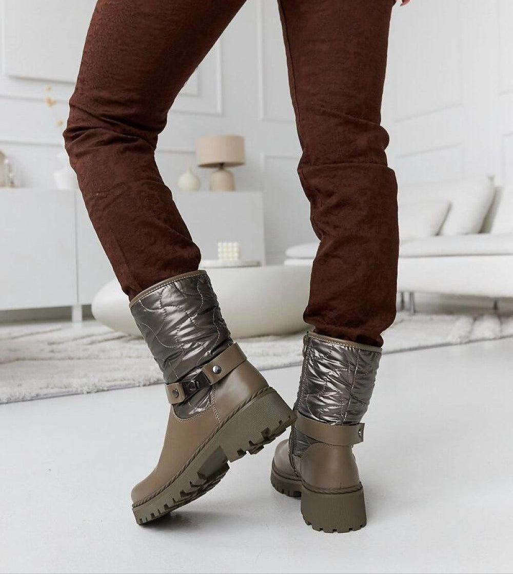 Brown Renvara strap detail ankle boots with decorative strap and side zipper, paired with dark brown pants in a living room setting.