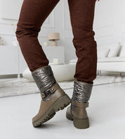 Brown Renvara strap detail ankle boots with decorative strap and side zipper, paired with dark brown pants in a living room setting.