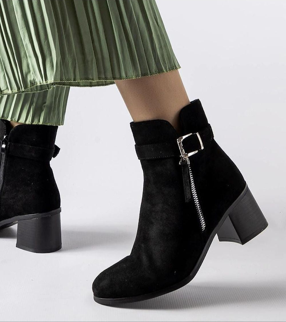 Esmyra low-heel ankle boots in black eco leather with zipper and buckle detail, paired with a green pleated skirt, side view.