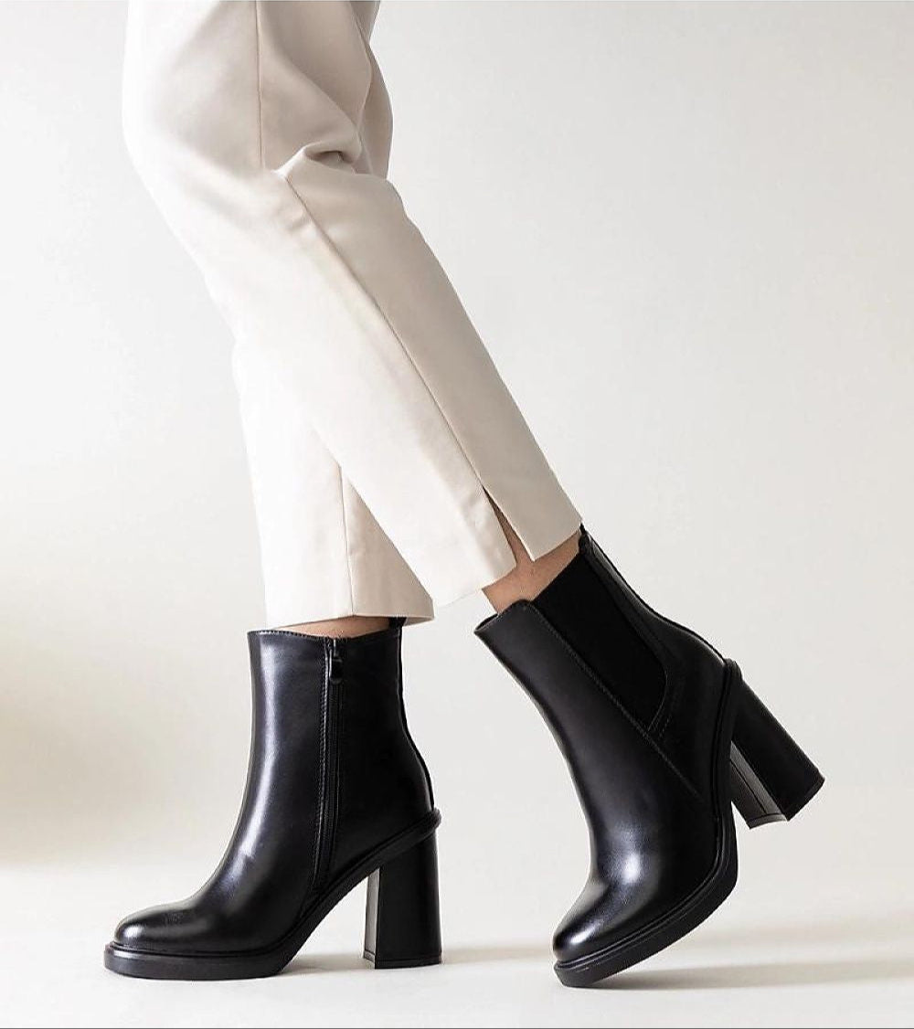 Woman wearing black stiletto zip-up boots with high heels and a sleek profile, paired with white pants, showcasing elegant footwear style.