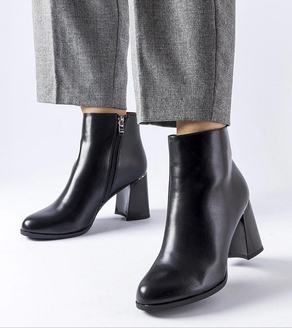 Black ankle boots with block heels and side zippers, paired with gray trousers, showcasing chic and modern footwear style.
