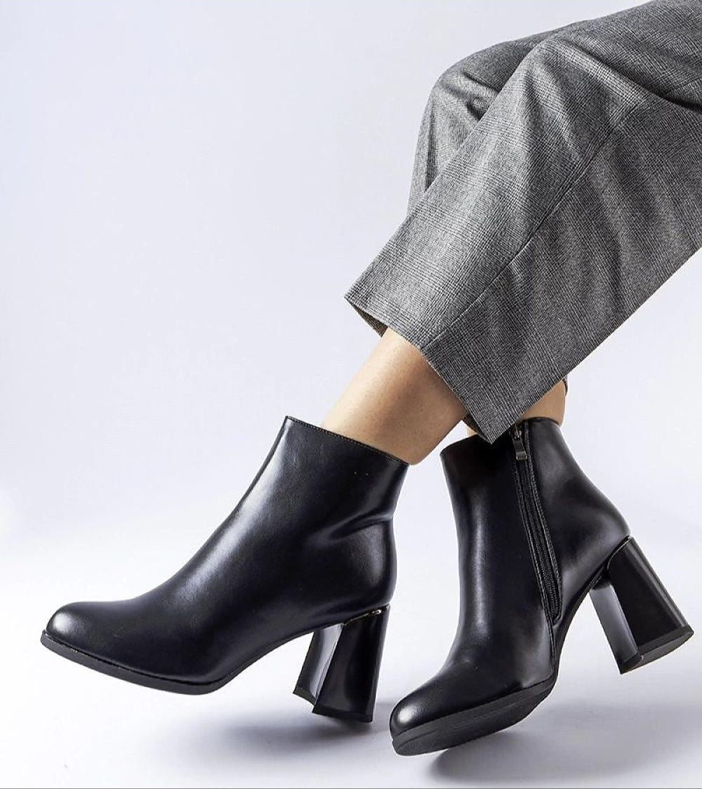 Person wearing black leather ankle boots with block heels and zipper closure, styled with gray trousers.