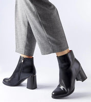 Person wearing black leather ankle boots with block heels and zipper, paired with gray plaid trousers.