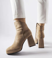 Stylish tan suede high-heeled ankle boots with white pants, featuring block heels and zipper closure on a white background.