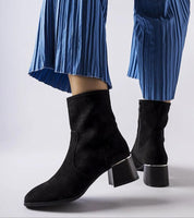 Black perforated zip-up ankle boots with a flat heel, styled with blue pleated pants for a chic, modern look.