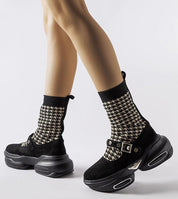 Person wearing black and white houndstooth knit socks with black platform sneakers and shiny buckles.