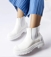 White slip-on bagger boots with rubber insets, featuring a platform sole and eco-suede material, paired with blue pleated pants.