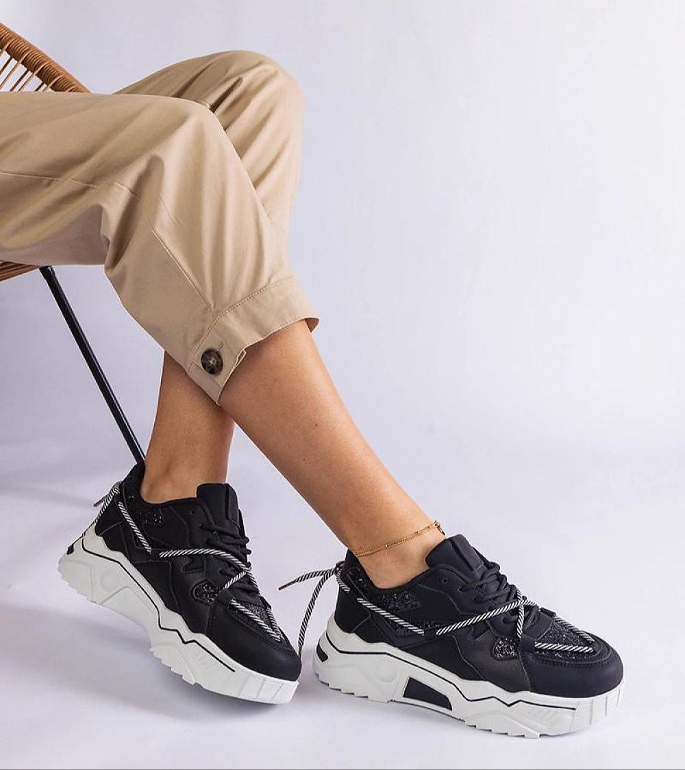 Person wearing Nimvara Shimmer Detail Black Sneakers with matte black brocade accents and white sole, styled with beige pants.