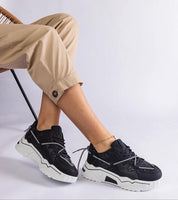 Person wearing Nimvara Shimmer Detail Black Sneakers with matte black brocade accents and white sole, styled with beige pants.