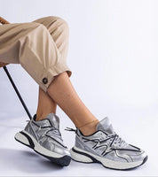Person wearing Yndessa Sculpted Sole Lace Sneakers in silver resting foot on chair, showcasing sleek design and comfort.
