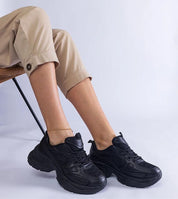 Person wearing black chunky sneakers with beige pants, seated on chair against white background.