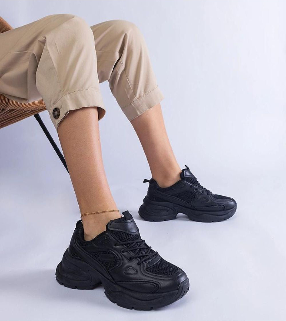 Person wearing black chunky sole sneakers with lace-up design and ankle-length beige pants, sitting on a chair.