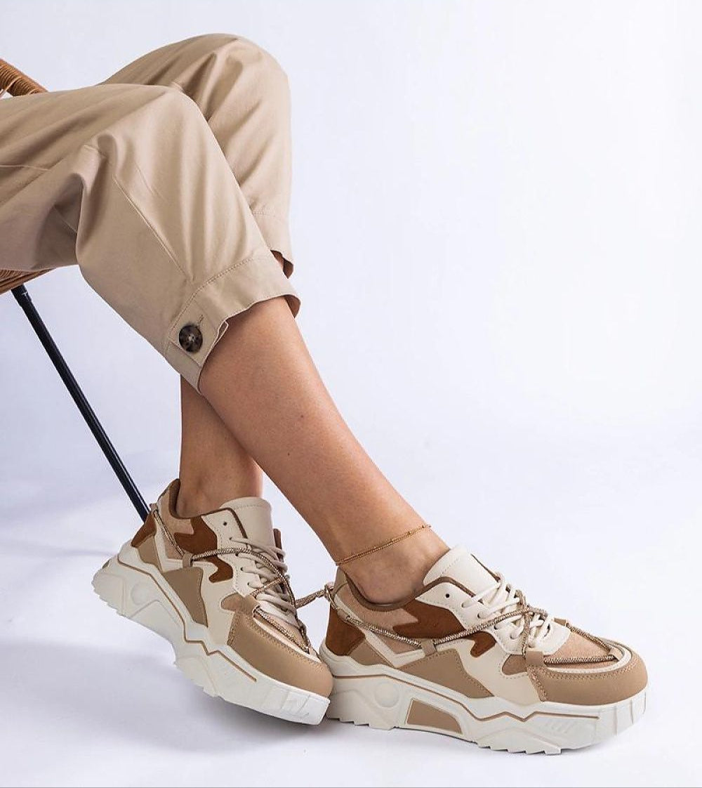 Person wearing beige and brown sneakers with a chunky white sole, styled with rolled-up khaki pants on a simple chair.