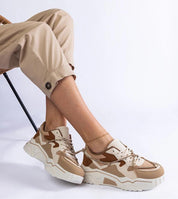 Person wearing beige and brown sneakers with a chunky white sole, styled with rolled-up khaki pants on a simple chair.
