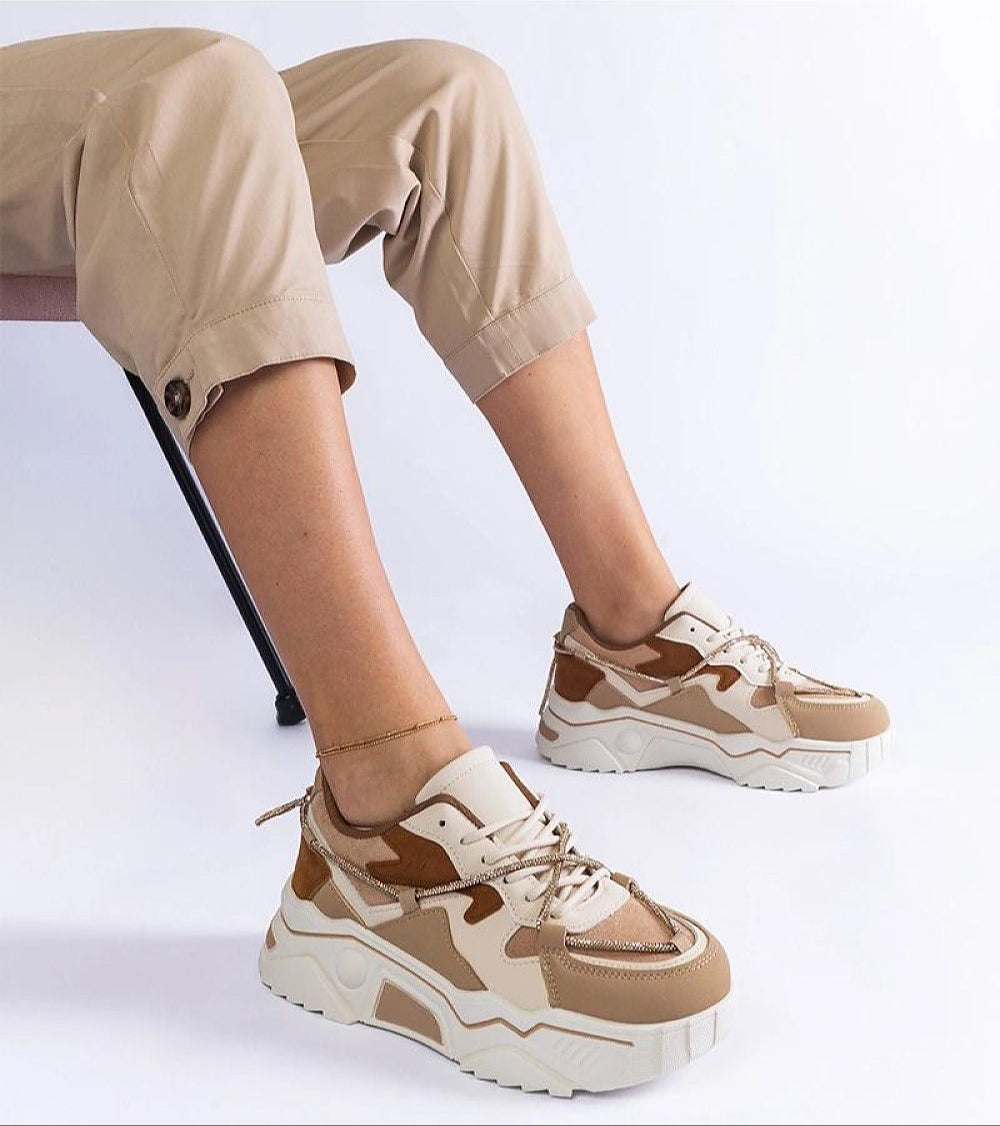 Person wearing Thalvyn Crystal-Tied Brown Sneakers with eco leather and crystal accents, sitting on chair with light beige trousers.