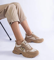 Person wearing Orevia Heart-Detailed brown sneakers with platform sole and heart accent, paired with beige pants, sitting in a chair.