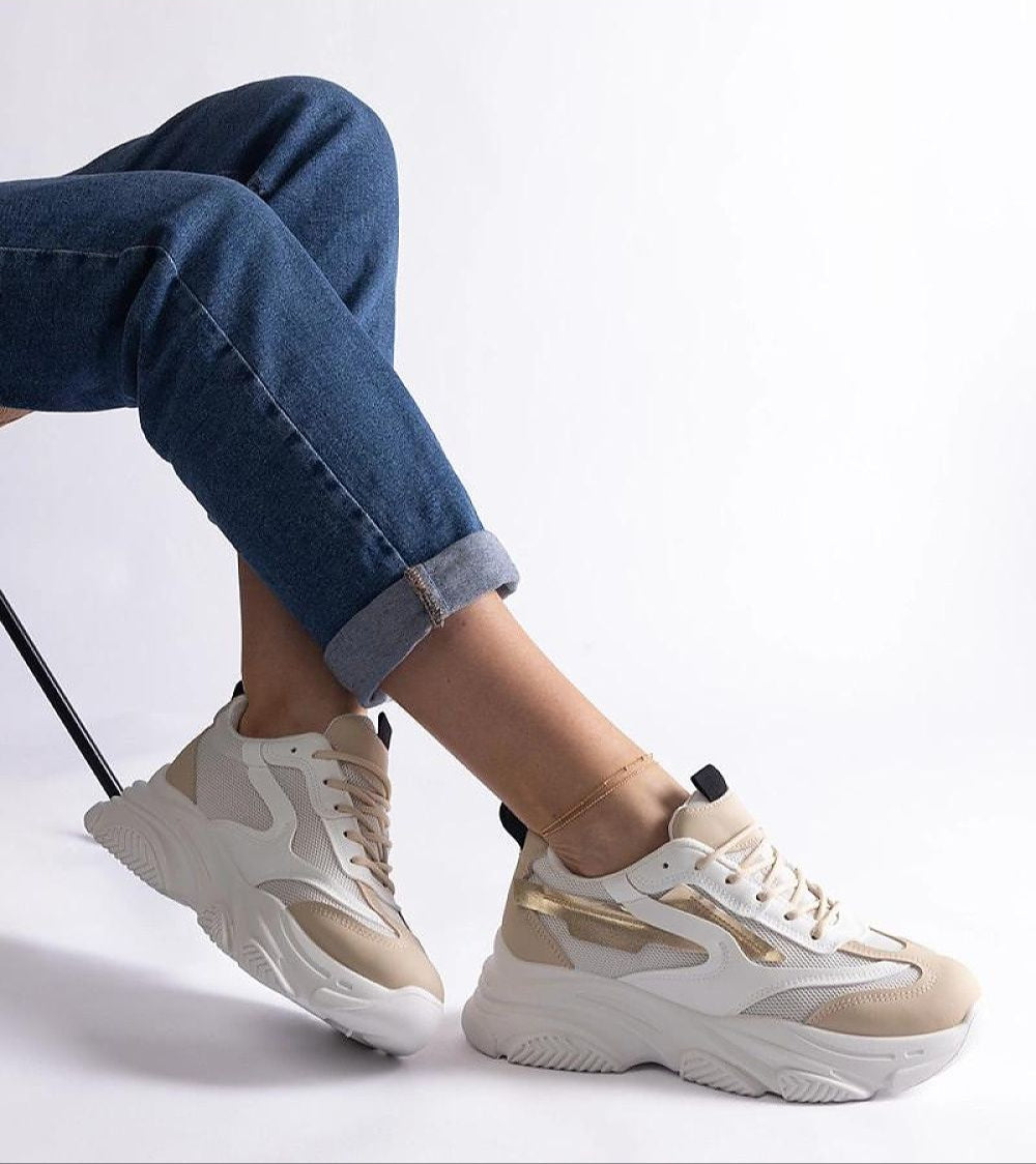 Stylish person wearing beige and white sneakers with thick sole, paired with rolled-up blue jeans, sitting on a chair against a white background.