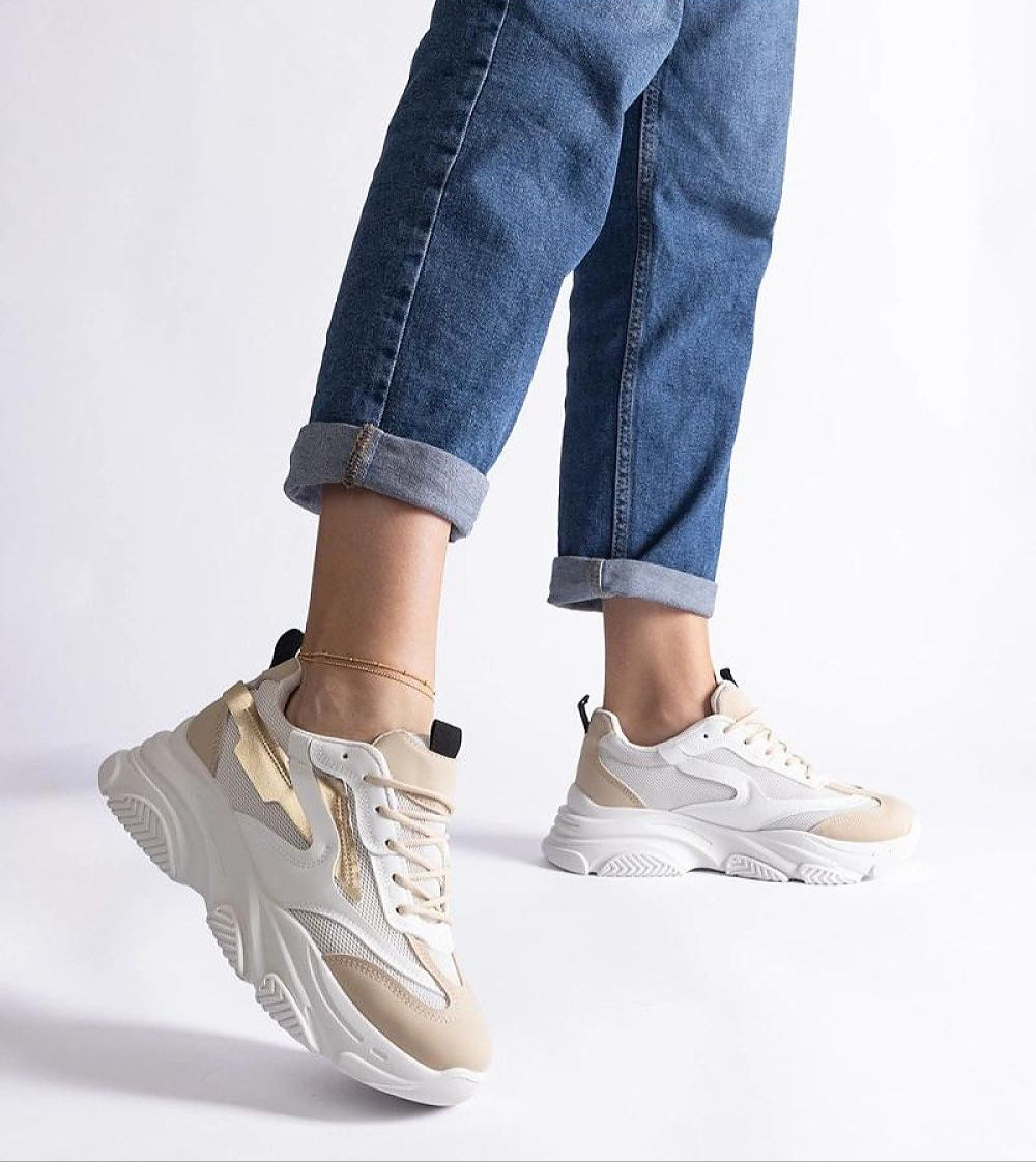 Person wearing beige and white chunky sneakers with blue jeans, showcasing casual urban footwear style on a white background.