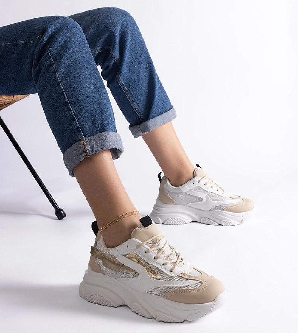 Woman wearing stylish beige-white sneakers with chunky sole and rolled-up jeans sitting on a chair against a white background.