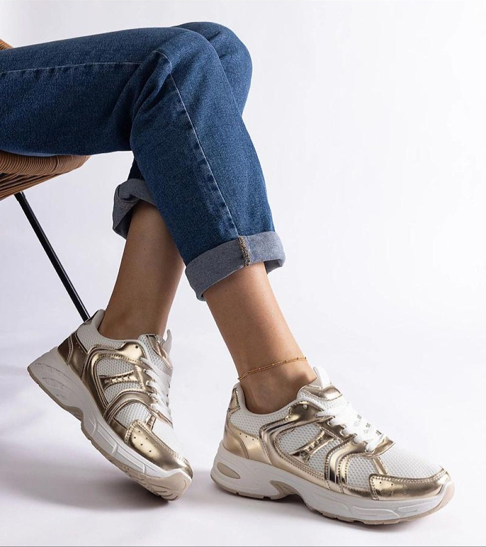 Woman wearing white and gold sneakers with denim jeans, seated stylishly on a chair, showcasing casual fashion.
