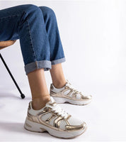 Person wearing white and gold sneakers with blue jeans, highlighting stylish footwear choice and casual outfit.