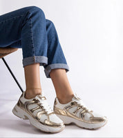 Person wearing stylish white and gold sneakers, paired with rolled-up denim jeans, seated casually on a chair.