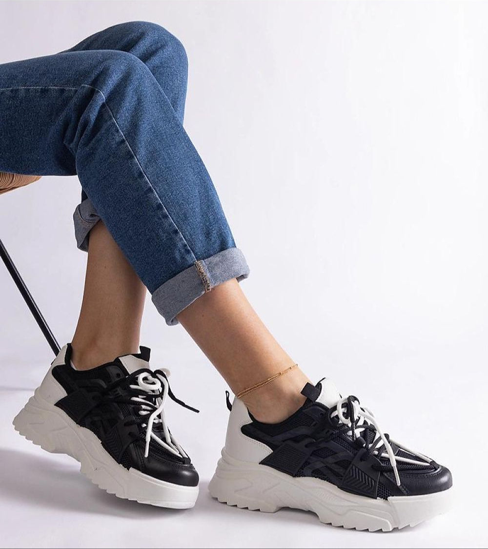 Person wearing black and white chunky sneakers with lace-up detail, paired with rolled-up blue jeans, sitting on a chair.