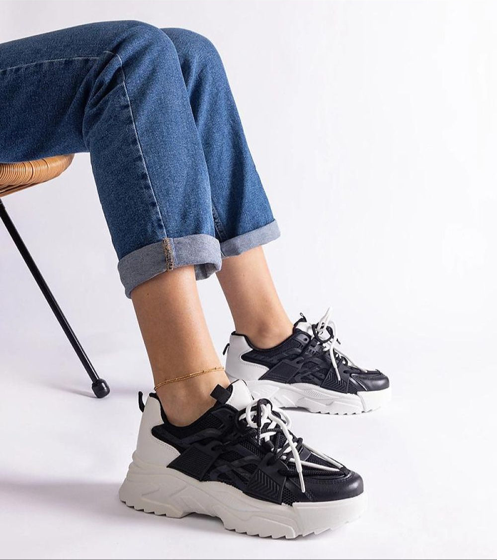 Person wearing black and white chunky sneakers with blue jeans, sitting on a chair.