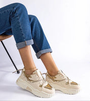 Person wearing white chunky sneakers with gold details and blue jeans, seated on a chair against a white background.
