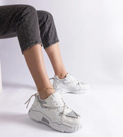 Person wearing white and silver sneakers with black jeans, ankle bracelet, seated on a white chair.